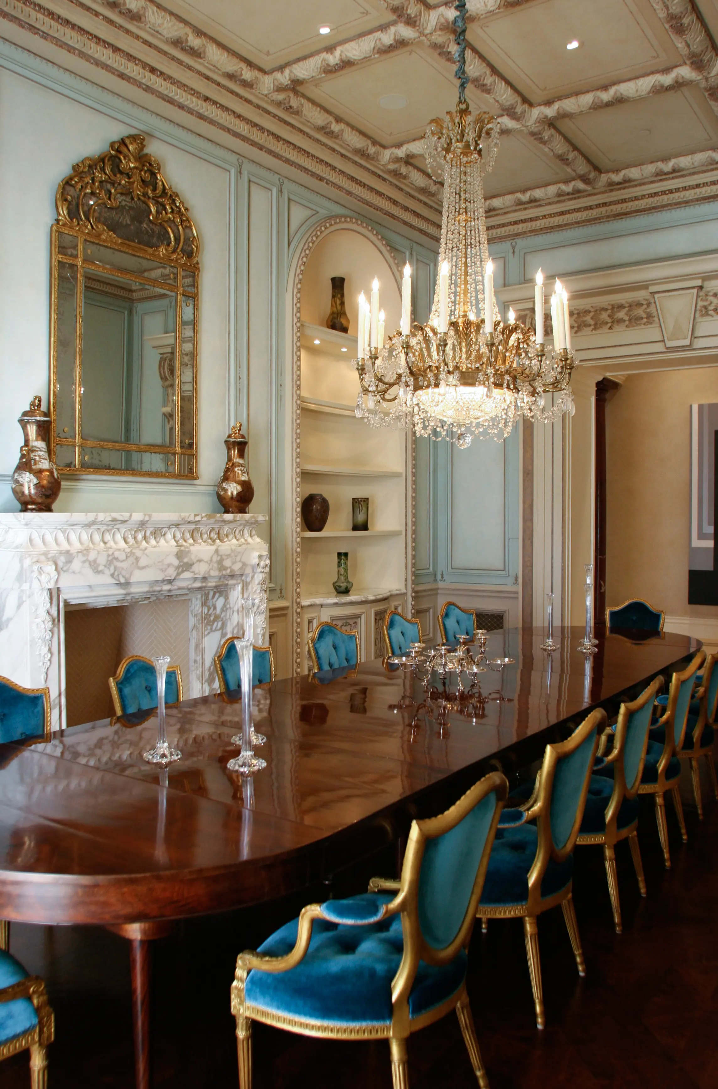 Formal dining room at the Merage House with pale blue glazed panelling, gilded mirror, carved marble chimneypiece and an ornamented coffered ceiling